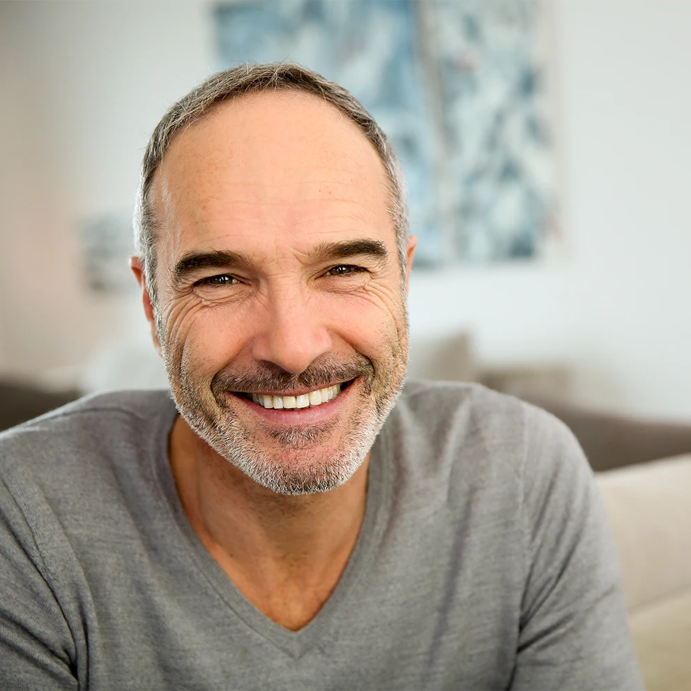 Smiling middle-aged man with short gray hair and a trimmed beard, wearing a light gray V-neck sweater. He has bright white teeth and slight crow’s feet, suggesting a genuine smile. The background shows a softly focused living room with a modern abstract painting.
