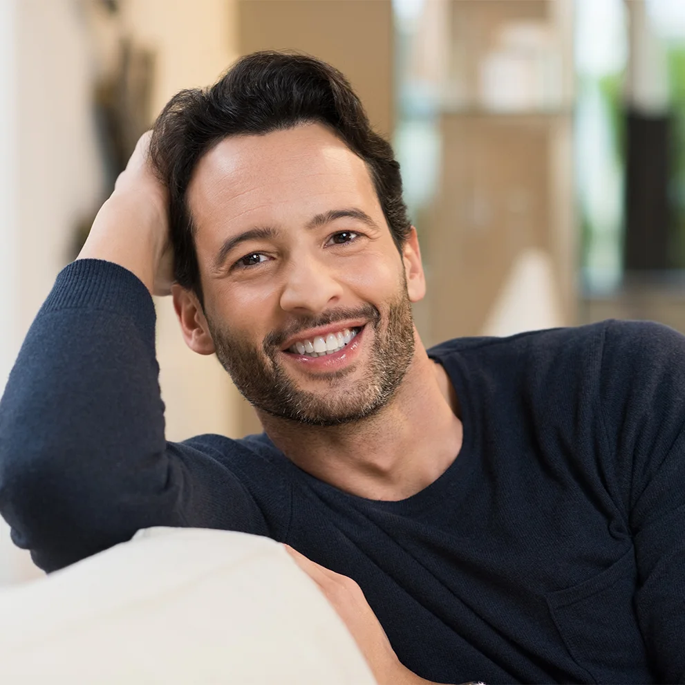 Smiling man with short dark hair and a trimmed beard, wearing a navy sweater and resting his head on his hand while leaning on a light-colored couch. His teeth are visible and well-maintained.