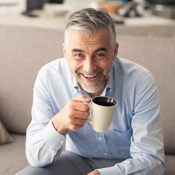 A mature man sitting on his couch with a cup of coffee while smiling at the camera