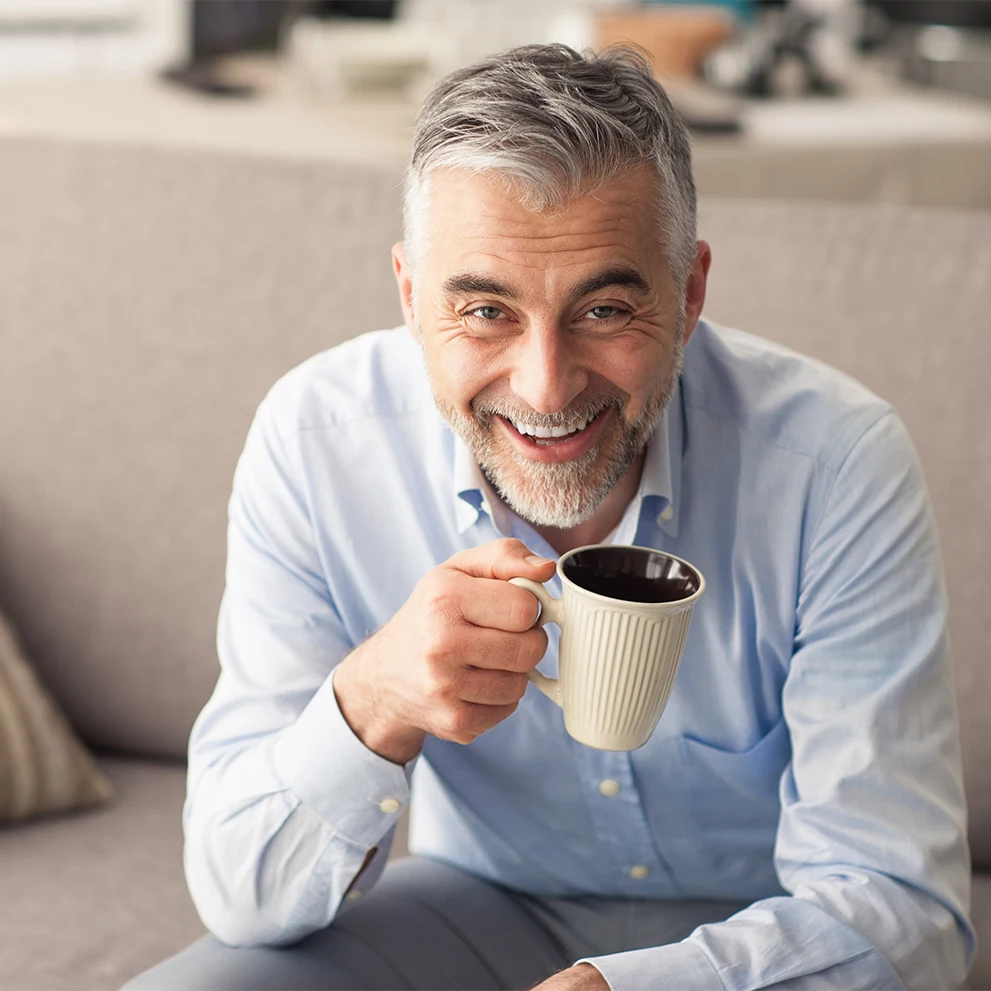 Smiling middle-aged man with gray hair and a neatly trimmed beard, holding a beige ceramic mug while sitting on a couch. He is wearing a light blue dress shirt and looking directly at the camera, with a warm, friendly expression. The background shows a softly focused kitchen area.