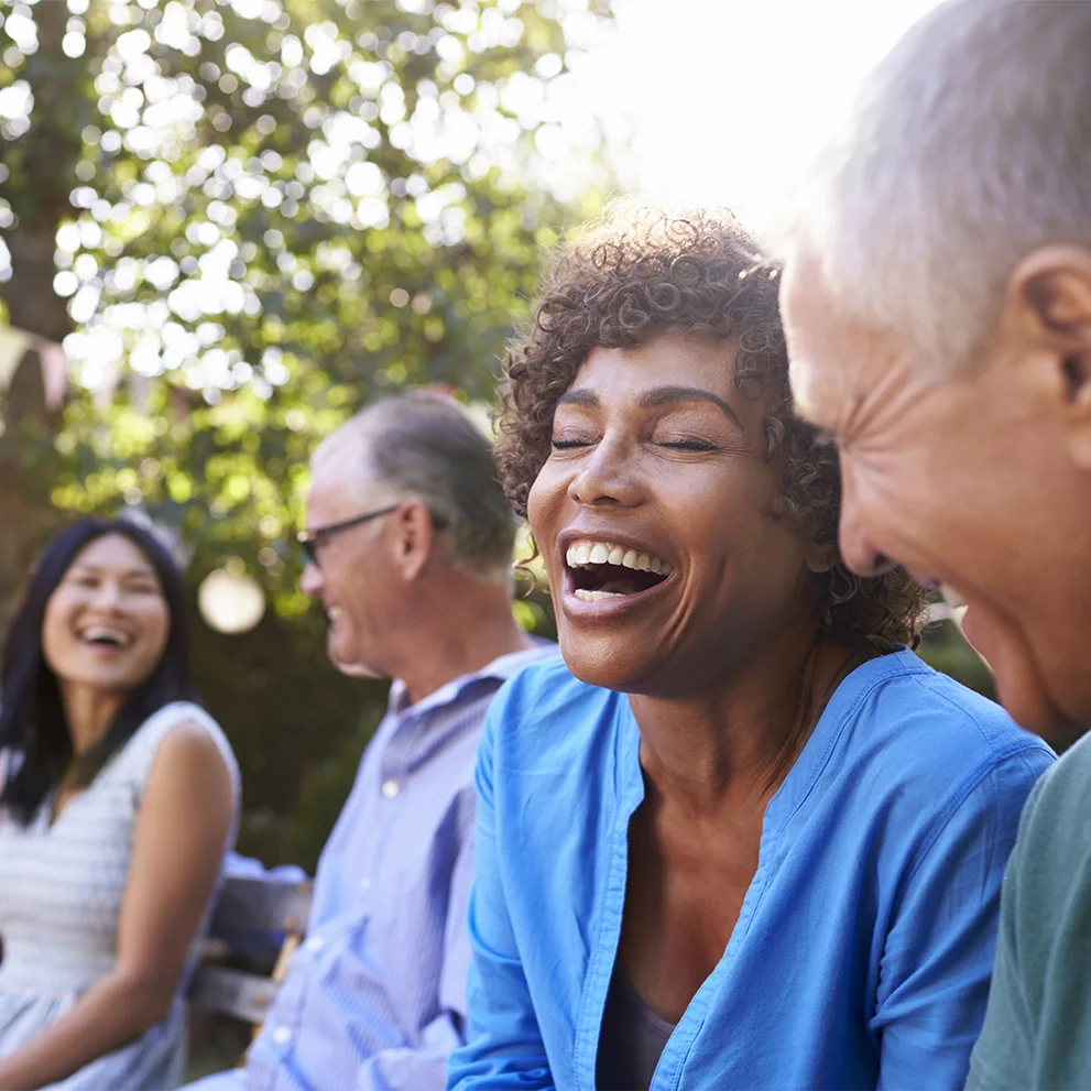 Group of four adults sitting on a bench outdoors, laughing and enjoying a sunny day. The woman in the foreground is wearing a blue top and smiling with her teeth visible, surrounded by trees and soft sunlight.