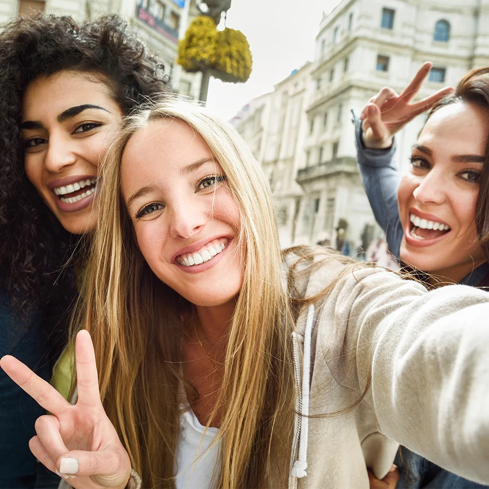 Three smiling young women posing for a selfie outdoors in an urban setting, all showing healthy, white teeth; suggests themes of dental aesthetics, oral hygiene, and confidence in one’s smile.