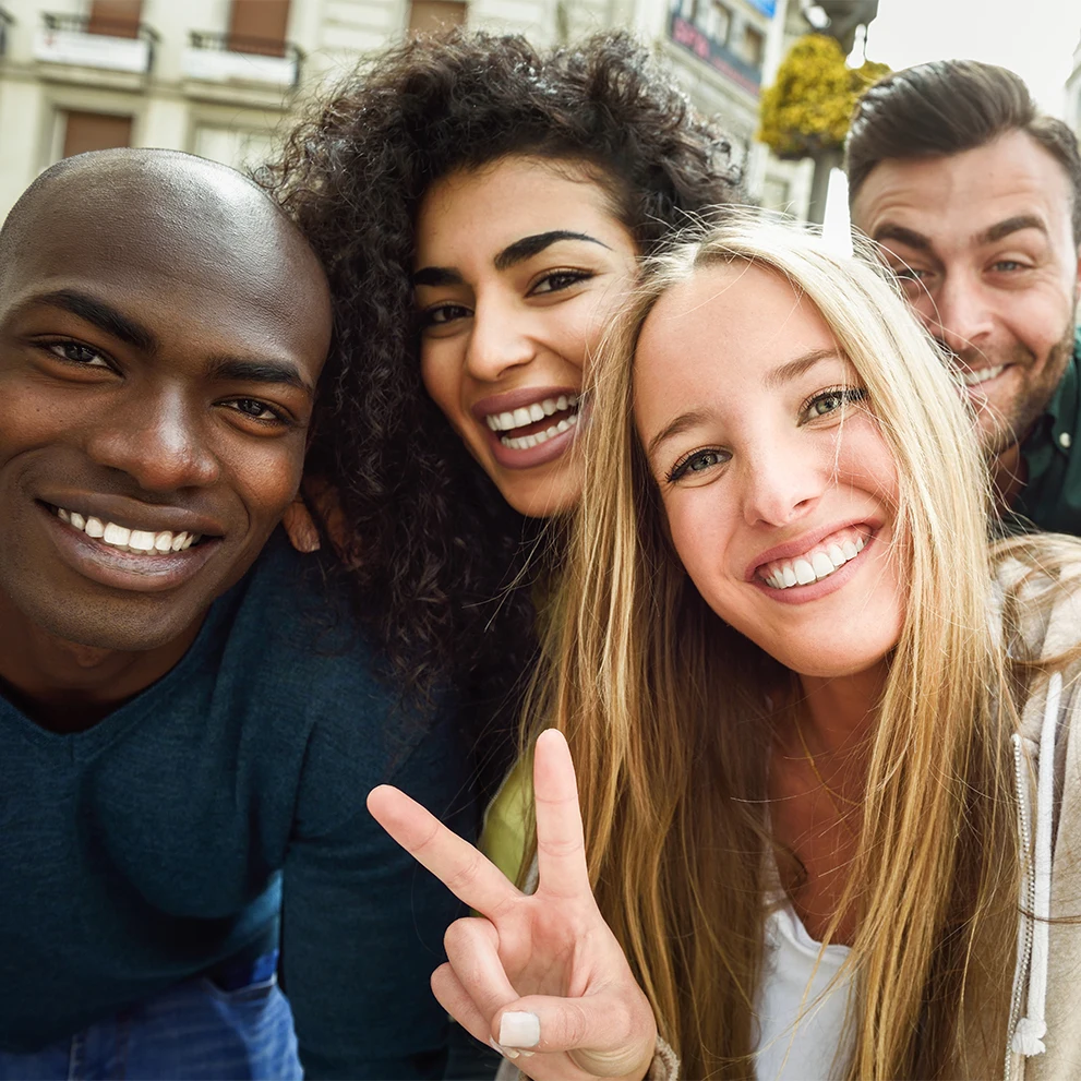 Four smiling friends posing closely together for a selfie outdoors in an urban setting. The woman in front with long blonde hair is holding up a peace sign. All four individuals are showing their teeth, with bright, well-aligned smiles. The background includes buildings and a tree-lined street.