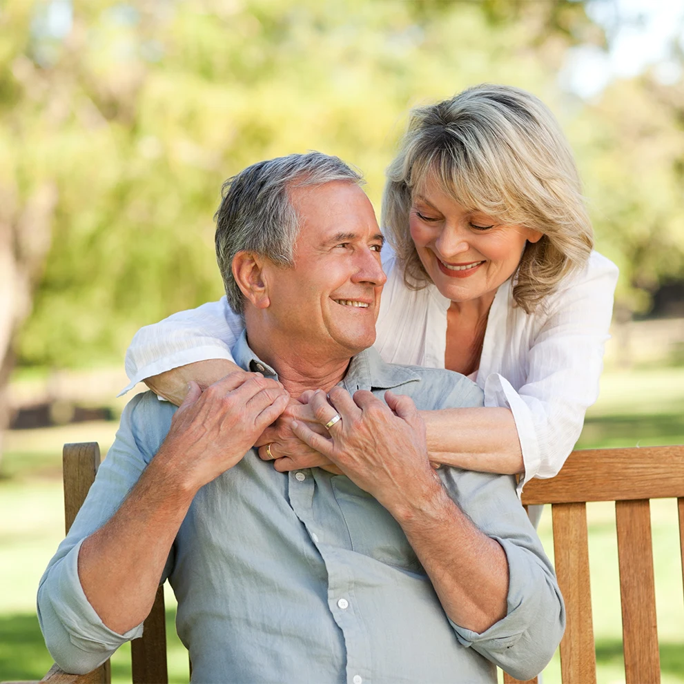 Smiling older couple sitting on a wooden bench in a park, with the woman leaning over from behind and embracing the man. Both are wearing light-colored clothing and wedding rings. They appear happy and relaxed, surrounded by greenery and soft natural light.