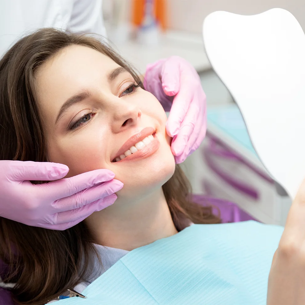Woman sitting in a dental chair smiling while looking in a tooth-shaped mirror. A dental professional wearing pink gloves is gently holding her face to show her results, and she is wearing a blue dental bib.