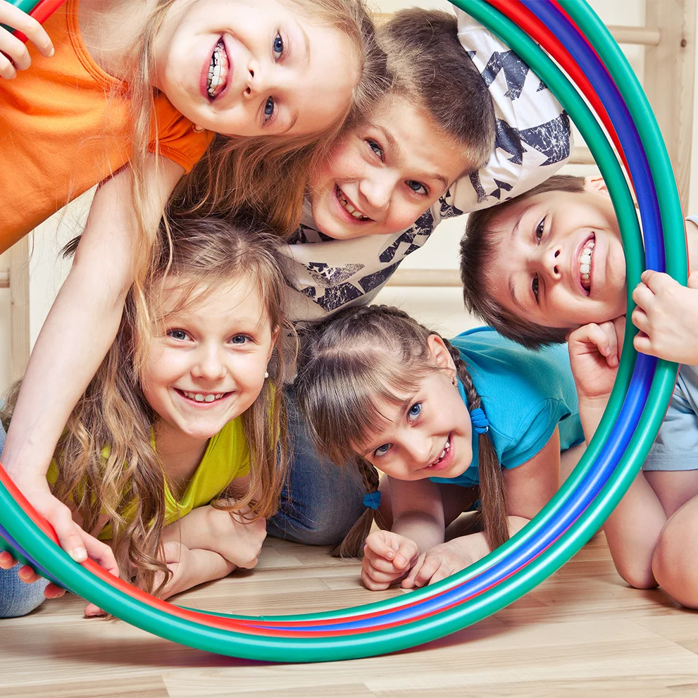 Group of five smiling children gathered closely on the floor, looking through colorful hula hoops. They are wearing bright clothing and appear cheerful and energetic.