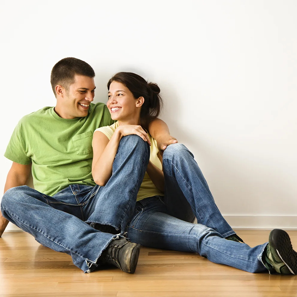 Smiling couple sitting on a wooden floor against a white wall, dressed casually in jeans and t-shirts. The man has his arm around the woman, and they are looking at each other warmly.
