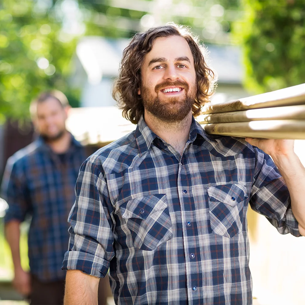 Smiling man with curly hair and a beard wearing a plaid shirt, carrying stacked wooden boards on his shoulder. Another man in a similar shirt is visible in the background. The scene is outdoors with bright natural light.