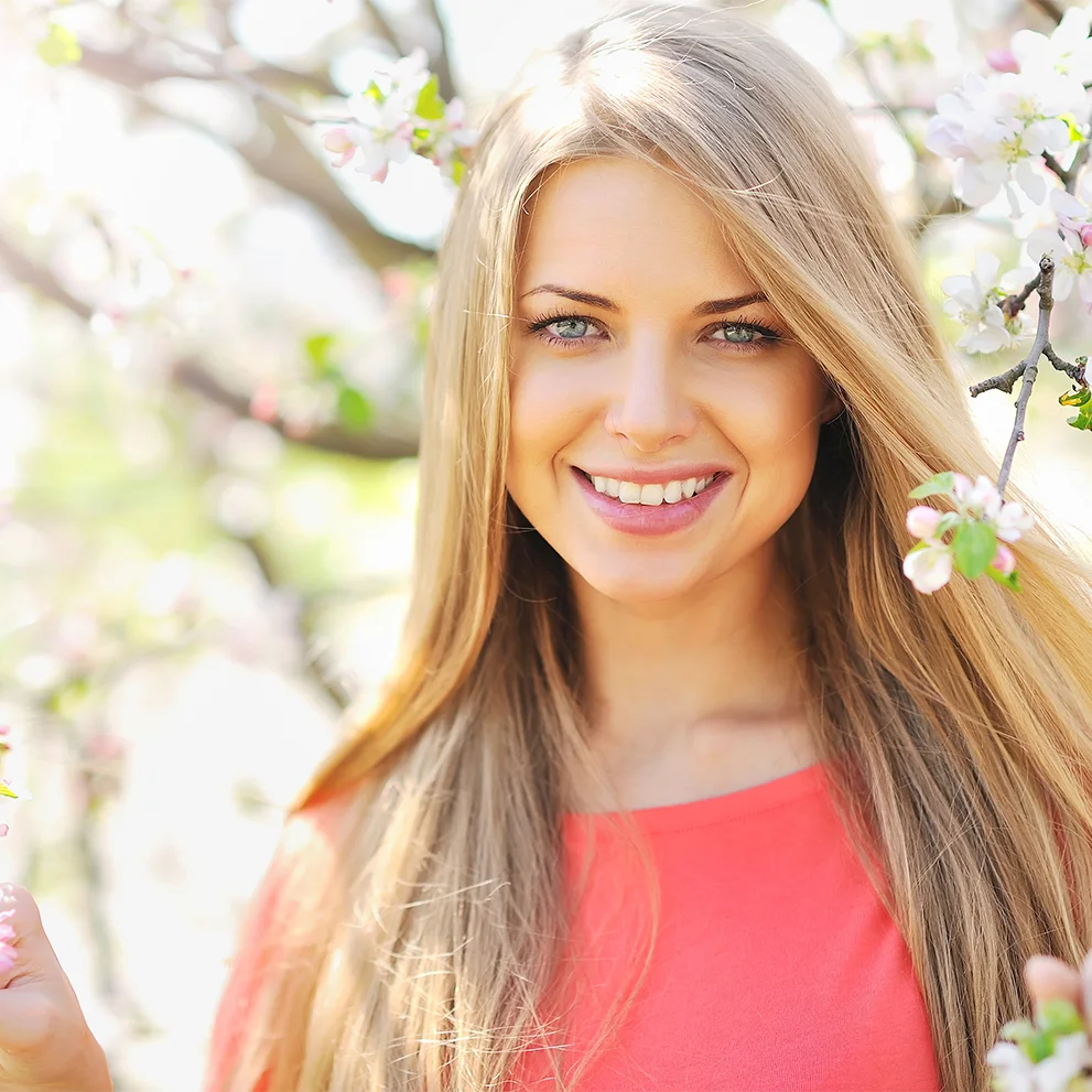Smiling young woman with long blonde hair wearing a coral top, standing among blooming tree branches in a sunlit outdoor setting. Her teeth are straight and white.