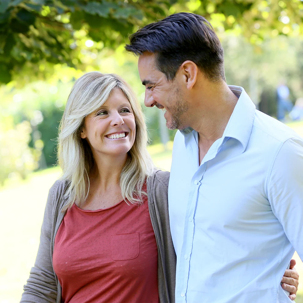Smiling couple walking outdoors under leafy trees, looking at each other affectionately. The woman has blonde hair and is wearing a red shirt with a brown cardigan, while the man has dark hair and is wearing a light blue button-down shirt. Sunlight filters through the greenery in the background.