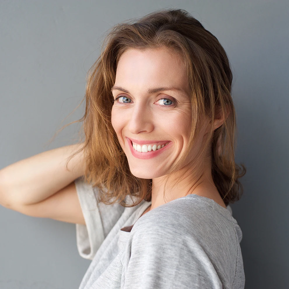 Smiling woman with wavy light brown hair wearing a light gray shirt, posing in front of a plain gray background. She has bright eyes, clear skin, and straight white teeth, with one arm raised behind her head.