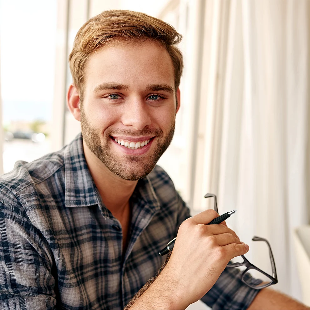 Smiling young man with neatly groomed beard and short light brown hair, wearing a plaid shirt and holding eyeglasses and a pen. He is seated indoors near a window with soft natural light illuminating his face, showing straight white teeth.
