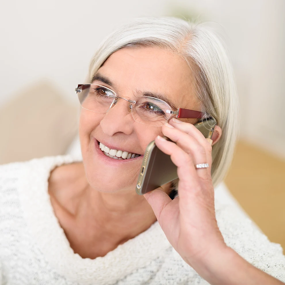 Smiling older woman with gray hair and glasses, holding a smartphone to her ear while talking. She is wearing a white textured sweater and a ring on her left hand. Her teeth are bright and well-aligned, and she appears happy and engaged in conversation.