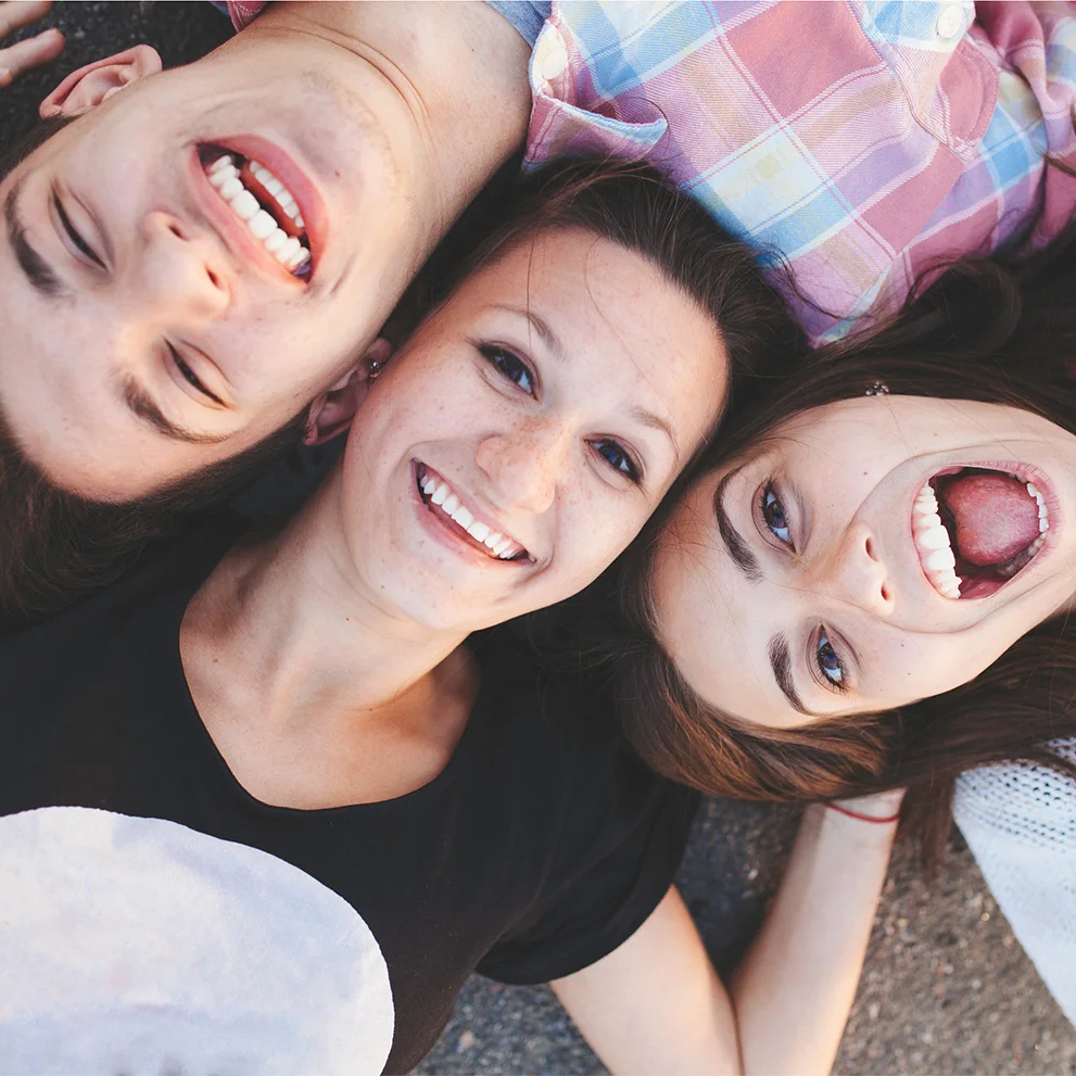 Three smiling friends lying on the ground in a circle with their heads together. All are showing their teeth, and the image is taken from above, capturing a playful and joyful moment.