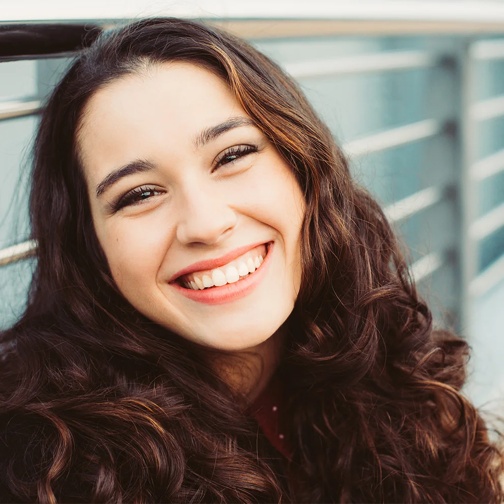 Close-up of smiling young woman with long curly brown hair and bright lipstick, leaning on a metal railing. Her teeth are well-aligned and she is outdoors in natural light.