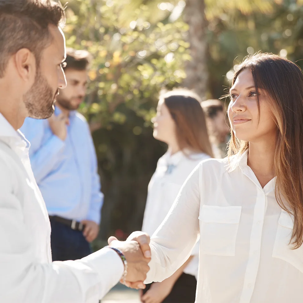 Man and woman shaking hands outdoors, both wearing white shirts. They are making eye contact with slight smiles, while two other people converse in the background among trees and sunlight.
