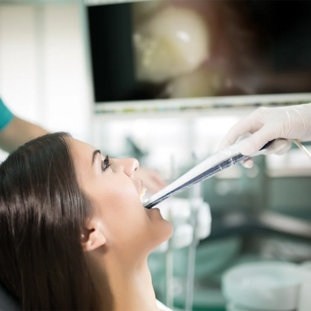 Patient in a dental chair undergoing an intraoral scan. A gloved hand holds an intraoral scanner inside the patient's open mouth, with a dental image visible on a screen in the background.