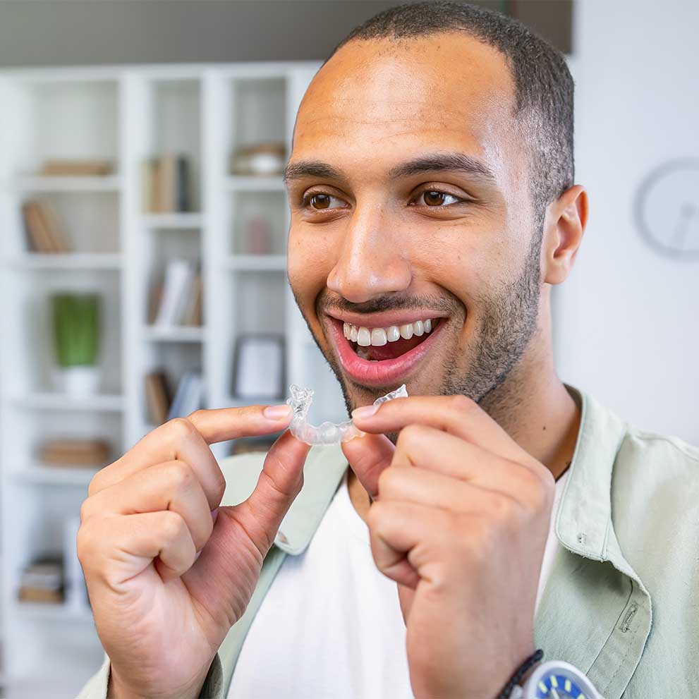 Smiling man holding a clear Invisalign aligner, preparing to place it over his upper teeth as therapy for teeth straightening.