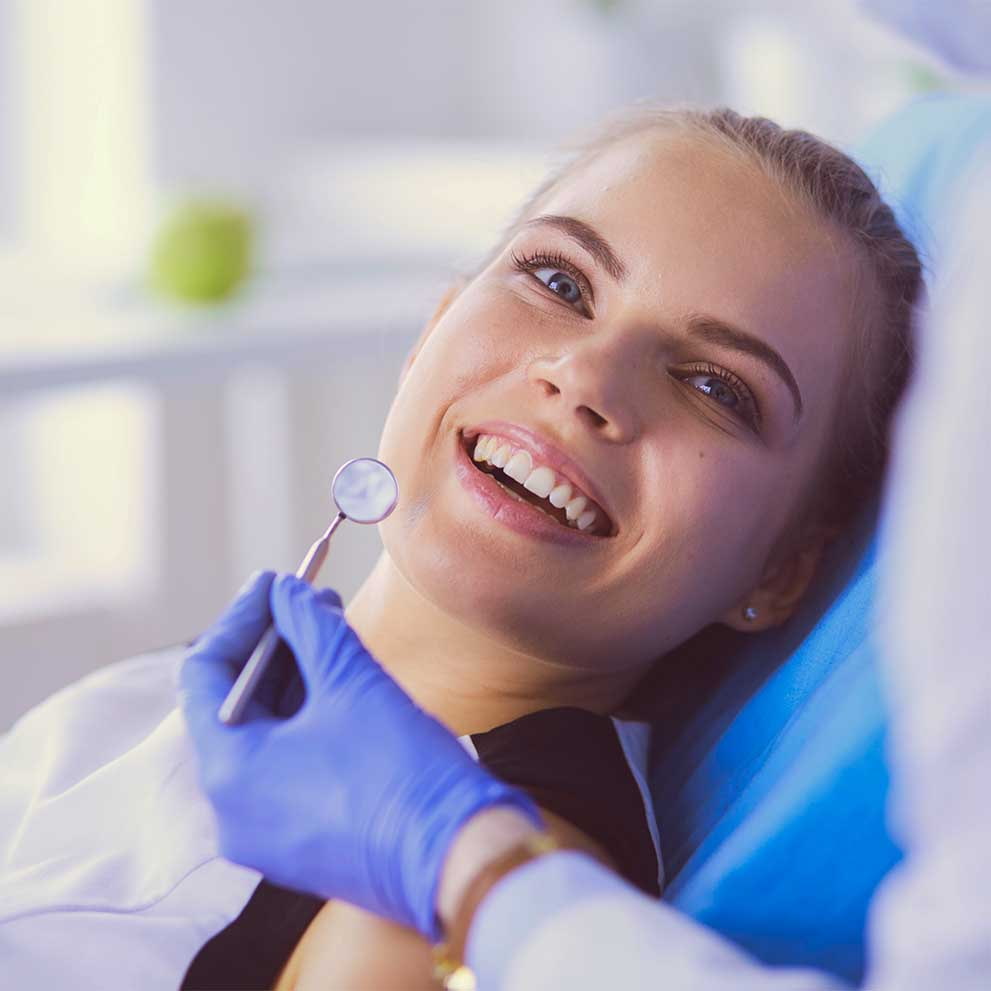 Smiling female patient reclining in a dental chair while a dentist wearing blue gloves holds a dental mirror near her mouth during an oral examination.